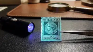 Francobollo italiano 20 lire Siracusana illuminato con lampada UV e pinzetta su piano da collezionista