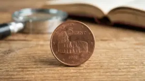 Moneta da 5 centesimi euro con il Colosseo in primo piano su un tavolo di legno, con lente d’ingrandimento sullo sfondo