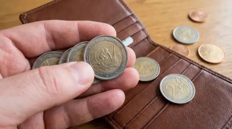 Mano che mostra una moneta da 2 euro commemorativa accanto a un portafoglio con altre monete su un tavolo