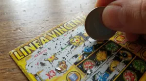 Mano che gratta un biglietto gratta e vinci con una moneta, con simboli di gioco visibili sul tavolo