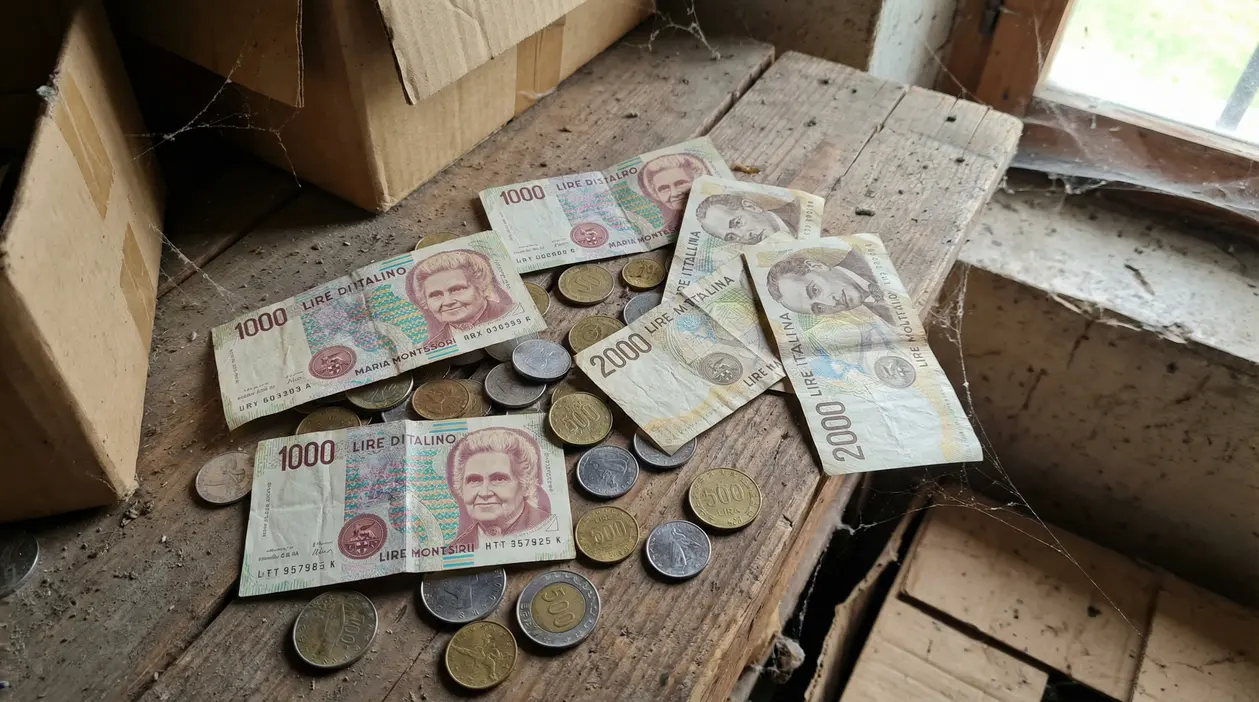 Banconote e monete in lire italiane trovate su un tavolo polveroso in soffitta, vicino a scatole di cartone