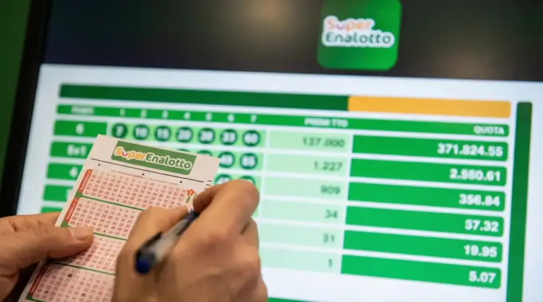 Mani che compilano una schedina del SuperEnalotto davanti a uno schermo con dati di quote e premi