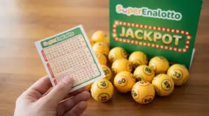 Mano che tiene una schedina del SuperEnalotto accanto a palline numerate e cartello del jackpot su un tavolo